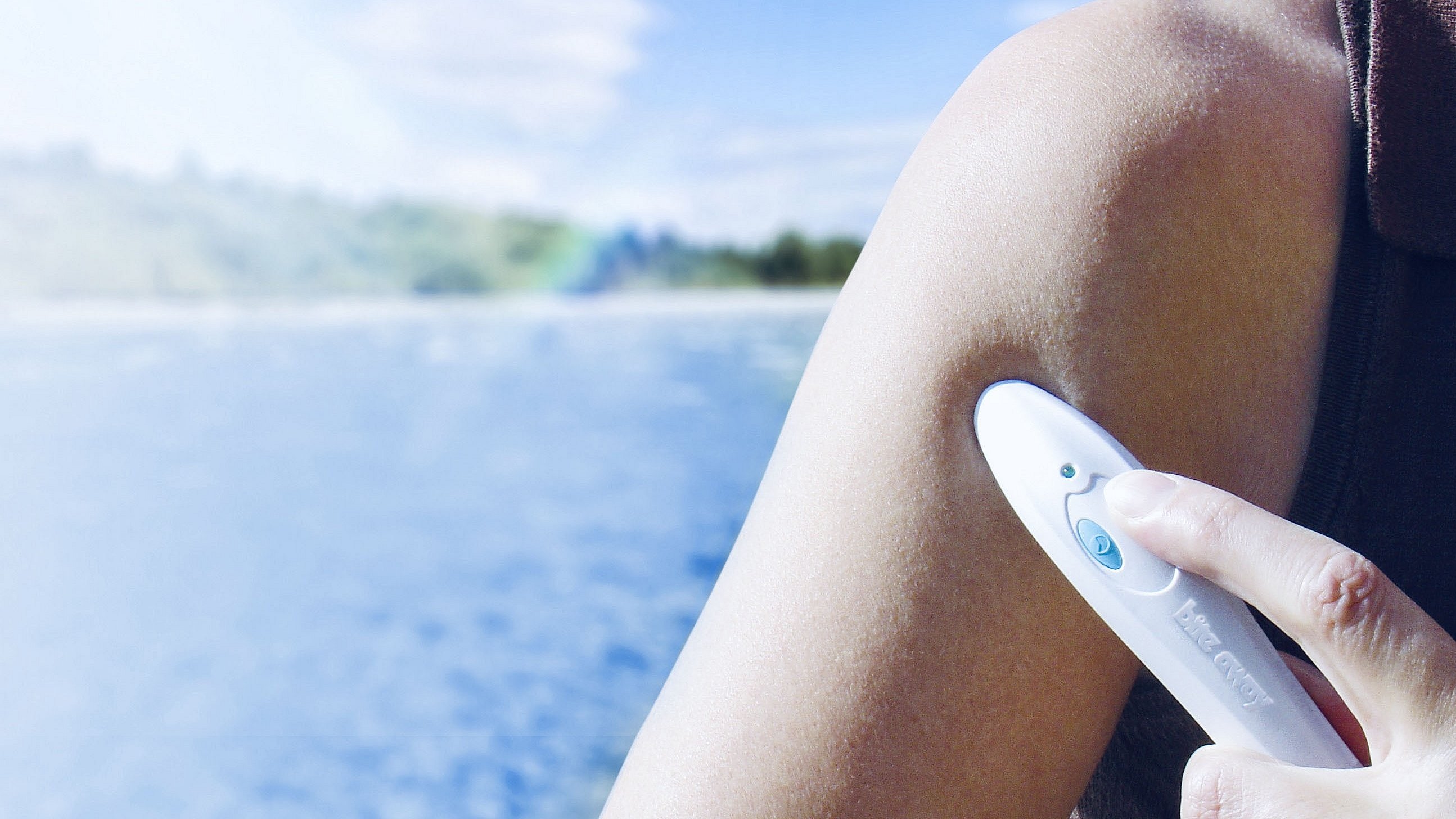 Eine Person wendet ein Bite-away Stichheiler-Gerät auf den Oberarm an. Im Hintergrund ist eine verschwommene Landschaft mit einem Gewässer und einem blauen Himmel zu sehen. Das Gerät ist weiß und hat einen blauen Knopf, der gedrückt wird.
