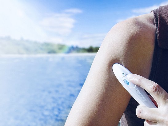 Eine Person wendet ein Bite-away Stichheiler-Gerät auf den Oberarm an. Im Hintergrund ist eine verschwommene Landschaft mit einem Gewässer und einem blauen Himmel zu sehen. Das Gerät ist weiß und hat einen blauen Knopf, der gedrückt wird.